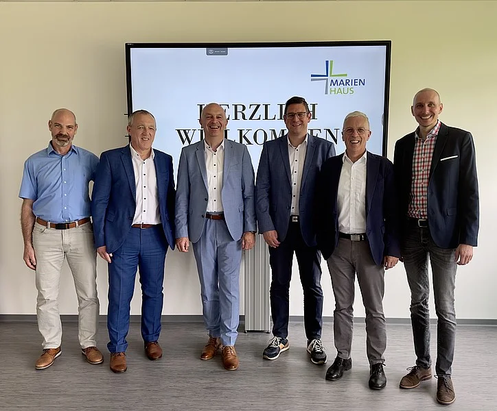 Sechs Männer in Anzügen und Hemden stehen vor einem Bildschirm mit dem Text 'HERZLICH WILLKOMMEN' und dem Logo von 'MARIENHAUS'.