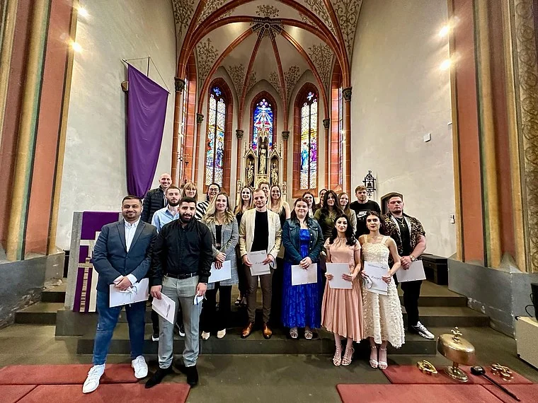Gruppe von Personen in formeller Kleidung, die in einer Kirche stehen. Im Hintergrund sind bunte Fenster und religiöse Statuen sichtbar. Einige halten Dokumente.