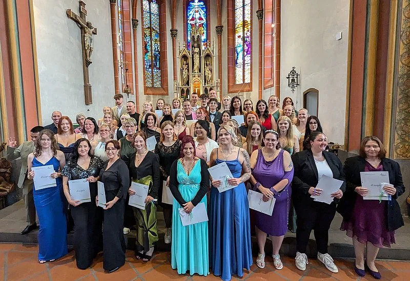 Gruppenfoto von 24 Personen in formeller Kleidung, die Urkunden halten, in einer Kirche mit bunten Glasfenstern im Hintergrund.